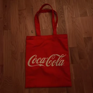 Röd Coca-Cola tote - Säljer en röd shoppingväska med Coca-Cola-logga. Perfekt för att bära med sig allt man behöver. Väskan har långa handtag som gör den enkel att slänga över axeln. En riktig klassiker för alla Coca-Cola-fans! 🥤