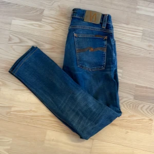 Blå jeans från nudie  - Snygga blå jeans med klassisk femficksdesign och orange sömmar. De har en knappgylf och en läderpatch baktill. Perfekta för en avslappnad stil.