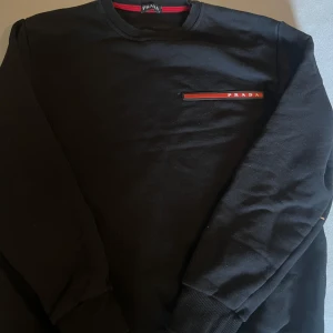 Svart tröja från Prada - Säljer en stilren svart tröja från Prada med en röd och vit detalj på bröstet. Tröjan har långa ärmar och en rund halsringning. Perfekt för en minimalistisk look.