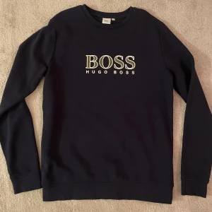 Svart tröja från Hugo Boss - Snygg svart tröja från Hugo Boss med deras ikoniska logga i gult på bröstet. Tröjan har långa ärmar och en klassisk rund halsringning. Perfekt för en stilren och avslappnad look.