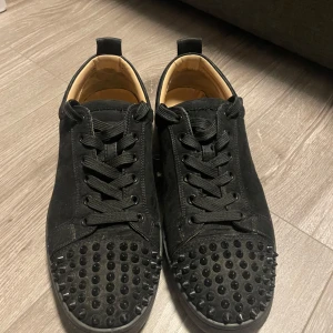 Louboutins med nitar  - Snygga svarta sneakers med nitar på tån och klassisk röd sula. Skorna har snörning och är tillverkade i mocka. Perfekta för att ge en edgy touch till din outfit.