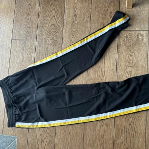 Svarta byxor från Gina Tricot - Snygga svarta byxor från Gina Tricot med gula och vita ränder längs sidorna. Byxorna har en elastisk midja och för extra komfort och är perfekta för en aktiv livsstil.