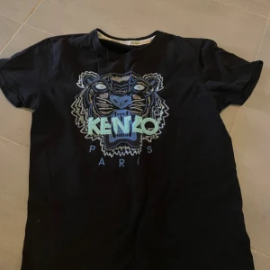 Svart t-shirt med tigerbroderi från Kenzo - Snygg svart t-shirt från Kenzo med ett stort, detaljerat tigerbroderi i flera färger på framsidan. T-shirten har korta ärmar och en rund halsringning. Perfekt för dig som vill ha en stilren men ändå iögonfallande look.