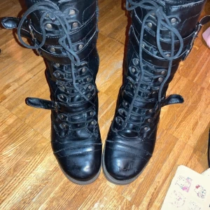 Svarta boots med snörning och spännen - Snygga svarta boots i skinn med snörning och dragkedja på sidan. De har även dekorativa spännen runt vristen och skaftet. Perfekta för en edgy look.  