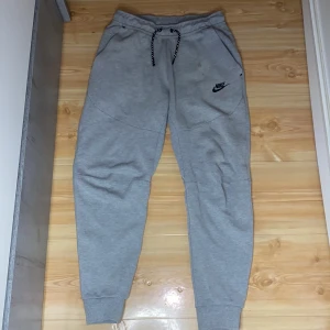 Grå mjukisbyxor från Nike - Säljer ett par grå mjukisbyxor från Nike med snörning i midjan och elastiska muddar vid bensluten. Byxorna har en liten Nike-logga på ena benet och är perfekta för en avslappnad stil.