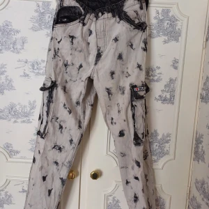 Grå och svarta cargobyxor - Unika grå cargobyxor med svart mönster och flera fickor. Byxorna har en loose passform och är perfekta för en avslappnad stil. De har knappar och dragkedja framtill samt justerbara band vid midjan och snören längst ner. Super coola men tyvärr förstora 