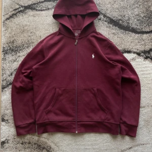 Vinröd hoodie från Polo Ralph Lauren - Säljer en snygg vinröd hoodie från Polo Ralph Lauren. Den har en dragkedja framtill och en klassisk logga på bröstet. Perfekt för en avslappnad stil.