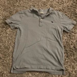 Ljusblå pikétröja kortärmad med knappar  - Säljer en ljusblå pikétröja från H&M med korta ärmar och en liten broderad detalj på bröstet. Tröjan har en klassisk krage med knappar framtill. Perfekt för en avslappnad stil.