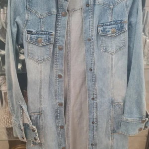 Blå lång jeansjacka från Crocker - Säljer en snygg blå jeansjacka från Crocker med knappar framtill och två bröstfickor med lock. Jackan har en klassisk design med kontrastsömmar och är perfekt för en avslappnad stil. Lång figursydd