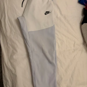 Ljusblå och vita Nike tech - Säljer ett par stilrena  nike tech  i en snygg kombination av ljusblått och vitt. Byxorna har en elastisk midja med svart snörning och Nike-logga på sidan. Perfekta för en avslappnad look. 147-158 cm