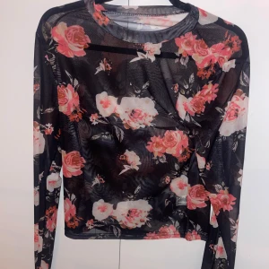 Svart blommig mesh-topp - Säljer en svart mesh-topp med vackert blommönster i rosa och vita nyanser. Toppen har långa ärmar och en rund halsringning, perfekt för att lägga till en romantisk touch till din outfit. Hund finns i hemmet.