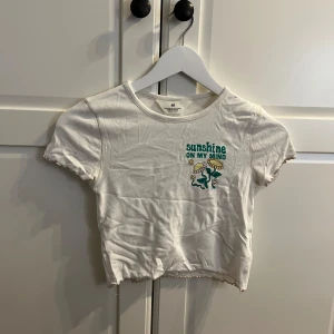Vit kortärmad topp från H&M - Säljer en vit kortärmad topp från H&M med trycket 'Sunshine on my mind' och en söt illustration framtill. Toppen är croppad och har en avslappnad passform. Perfekt för soliga dagar! ☀️