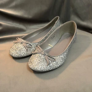 Glittriga ballerinaskor med rosett - Snygga glittriga ballerinaskor i silver med en söt rosett på tån. Perfekta för att lägga till lite bling till din outfit. Skorna har en bekväm passform och en dekorativ sula.