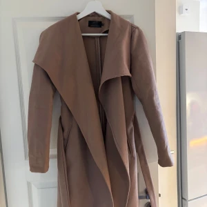 Brun/beige kappa med bälte - Snygg beige/brun  kappa med bälte och långa ärmar. Perfekt för höst och vår, med en stilren design som passar till många tillfällen. Materialet ser mjukt och bekvämt ut, vilket gör den till ett mångsidigt plagg i garderoben. Finns två små fläckar som säkert går bort men jag har inte försökt. Annars väldigt fint skick! 