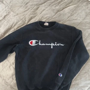 Svart sweatshirt från Champion - Säljer en svart sweatshirt från Champion med det ikoniska logotyptrycket i vitt och rött på bröstet. Tröjan har långa ärmar och en rund halsringning. Perfekt för en avslappnad stil.
