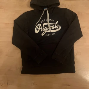 Svart hoodie från Jack & Jones - Säljer en svart hoodie från Jack & Jones med vit text på framsidan. justerbar huva med snören. Perfekt för en avslappnad stil.