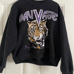 Svart sweatshirt med tigertryck från Stay - Cool svart sweatshirt från Stay med ett stort tigertryck och texten 'Sauvage' i lila och vitt på framsidan. Tröjan har en avslappnad passform och långa ärmar, perfekt för en casual look.