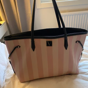 Victorias Secret väska  - Snygg rymlig väska med breda rosa och vita ränder. Väskan har svarta handtag och detaljer, samt en praktisk dragkedja upptill. På insidan finns ett fack. Perfekt väska som du kan ha till skolan, jobbet eller var som helst! 