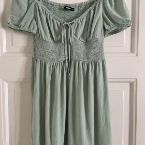Grön klänning från BikBok - Säljer en söt grön klänning från BikBok med korta puffärmar och smockdetaljer i midjan. Klänningen har en knytning framtill och en luftig passform som passar perfekt för varma dagar.