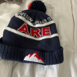 Mörkblå Åre mössa med tofs - Stickad mörkblå mössa med vit och röd text 'ÅRE' framtill, vita ränder och broderat bergmotiv på uppviket. Toppad med en fluffig tofs i rött och mörkblått. Perfekt för kalla dagar och vintersäsong.