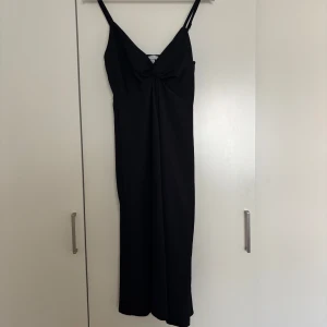 Svart långklänning med knut H&M - Stilren svart långklänning från H&M med smala axelband och snygg knutdetalj framtill. Klänningen har en elegant v-ringning och är tillverkad i ett lätt, strukturerat tyg som ger en lyxig känsla. Perfekt för dig som gillar minimalistisk och chic stil.