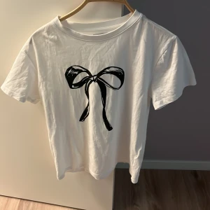 Vit t-shirt med svart rosett Holly & Whyte - Vit t-shirt från Holly & Whyte by Lindex med ett stort svart rosett-tryck på bröstet. Klassisk rund halsringning och korta ärmar. Tillverkad i mjuk bomull som känns skön mot huden. Perfekt för dig som gillar en söt men stilren look.