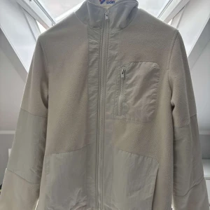 Beige fleecejacka med dragkedja - Snygg beige fleecejacka  dragkedja framtill och en bröstficka med dragkedja. Jackan har paneler i ett slätt material på bröst, ärmar och sidor för en cool kontrast. Perfekt för lager-på-lager och chill vardagsstil.