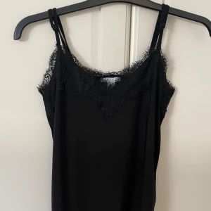 Svart linnetopp med spetsdetaljer - Snygg svart linnetopp med tunna axelband och vacker spets längs urringningen. Materialet känns mjukt och följsamt mot huden.