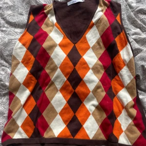 Rutig stickad väst - Snygg stickad väst från Almia i storlek XXL med rutigt mönster i brunt, orange, vinrött, beige och vitt. Västen har v-ringning och är ärmlös, perfekt att styla över en skjorta eller t-shirt för en cool look.