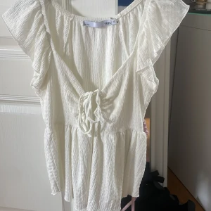 Vit blus med knyt och volang - Supersöt vit blus i crinklat tyg med v-ringning, knyt framtill och volangdetaljer vid axlarna. Lätt och luftig modell som passar perfekt till sommaren. Kortärmad och med avslappnad passform.