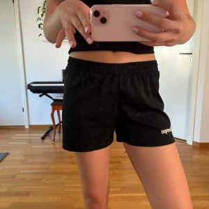 Svarta shorts från Kempa - Svarta sportiga shorts från Kempa med elastisk midja och kort passform. Perfekta för träning eller chill, med diskret logga på ena benet. Lätta och bekväma, tillverkade i mjukt syntetmaterial som andas.