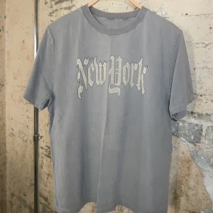 Grå t-shirt med New York-tryck - Snygg grå t-shirt med coolt New York-tryck i gotisk stil på bröstet. Klassisk passform med rund hals och korta ärmar. Perfekt för dig som gillar streetwear och vill ha en chill vibe.