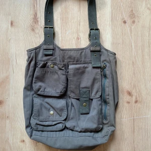 Grön axelväska med fickor - Rymlig axelväska i bomull med flera praktiska fickor framtill, både med tryckknappar och dragkedja. Justerbara axelband i grönt tyg och en stor öppen ficka på baksidan. Perfekt för dig som gillar funktionell och cool stil.