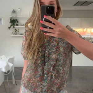 Blommig volangskjorta från Zara - Supersöt blommig skjorta från Zara med volangdetaljer framtill och knappar hela vägen. Skjortan är tunn och luftig med korta ärmar, perfekt för varmare dagar. Färgerna går i grönt, rosa och vitt och materialet känns lätt och somrigt.