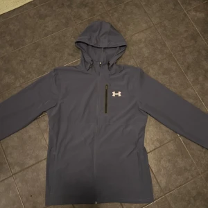 Mörkblå jacka från Under Armour - Säljer en mörkblå jacka från Under Armour med huva och dragkedja. Jackan har en bröstficka med dragkedja och det ikoniska Under Armour-logotypen på bröstet. Perfekt för en sportig look!