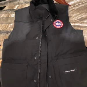 Svart dunväst från Canada Goose - Svart dunväst från Canada Goose med hög krage, två stora fickor framtill och klassisk logotyp på bröstet. Västen har dragkedja och tryckknappar samt diskret Canada Goose-märke på ena fickan. Perfekt för lager-på-lager under kyliga dagar.