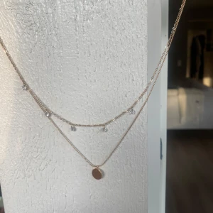 Guldigt dubbelt halsband med berlocker - Snyggt dubbelt halsband i guldton med tunn kedja. Det övre lagret har små glittrande stenar som hänger längs kedjan och det undre lagret har en rund, slät berlock. 