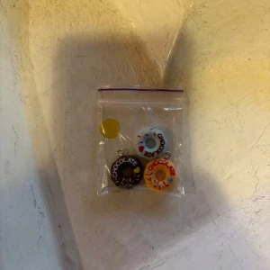 Donut-berlocker med färgglada detaljer - Tre söta örhängen formade som donuts i olika färger: chokladbrun, vit och gul. Alla har färgglada strössel och texten 'CHOCOLATE' på sig. Perfekta för dig som gillar roliga och unika accessoarer med lekfull stil.