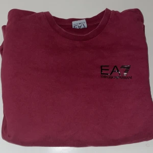 Vinröd tröja ifrån EA7 Armani - Vinröd långärmad tröja från EA7 Emporio Armani med svart logga på bröstet. Klassisk rund halsringning och mjukt bomullsmaterial som känns skönt mot huden. Perfekt för dig som gillar stilrena och snygga basplagg med premiumkänsla.