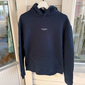 Mörkblå hoodie från Axel Arigato - Snygg mörkblå hoodie från Axel Arigato i 100% ekologisk bomull. Klassisk modell med huva, känguruficka och diskret vit logga på bröstet. Perfekt för dig som gillar stilrena och bekväma plagg med streetkänsla. Tillverkad i Portugal. Bra skick. 
