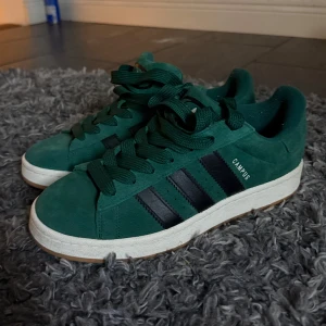 Adidas Campus gröna sneakers - Gröna Adidas Campus sneakers i mocka med svarta ränder på sidorna och vit mellansula. Klassisk låg modell med rund tå, gröna skosnören och gummisula i naturfärg. Perfekta för dig som gillar retrostil och streetwear. Bra skick använda 2-3 gånger bara. Storlek 44