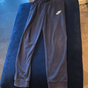 Mörkblå mjukisbyxor från Nike XL - Mörkblå mjukisbyxor från Nike i storlek XL, med klassisk logga på vänster höft. Byxorna har resår i midjan och muddar vid bensluten för en skön passform. Perfekta för chill eller träning, tillverkade i mjukt material. Jacka finns till i annan annons 