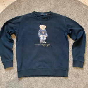 Mörkblå sweatshirt med Polo Bear från Ralph Lauren - Säljer en mörkblå sweatshirt från Ralph Lauren med det klassiska Polo Bear-motivet framtill. Tröjan har rund halsringning och långa ärmar. Perfekt för dig som gillar streetwear och ikoniska prints. Storlek M men passar mer S. Tröjan är bra skick andvänd mycket och därför är den blå färgen lite ut tvättad och logan är lite sliten 