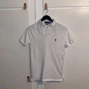Vit kortärmad pikétröja från Ralph Lauren - Snygg vit pikétröja från Ralph Lauren med klassisk krage och korta ärmar. Tröjan har en broderad logotyp på bröstet. Perfekt för en stilren och avslappnad look.