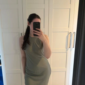 Olivgrön ärmlös långklänning - Säljer en stilren olivgrön långklänning med ärmlös design och rund halsringning. Klänningen har en rak passform och faller snyggt längs kroppen, perfekt för dig som gillar minimalistisk stil. Materialet känns mjukt och svalt mot huden.