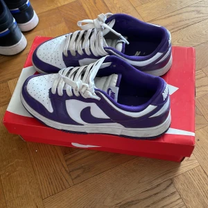 Nike Dunk Low Retro vit/lila - Nike Dunk Low Retro sneakers i vit och lila med klassisk låg siluett. Skorna har vit bas med lila overlays och Swoosh, samt matchande lila innerfoder. Ovandel i skinn och snörning framtill. Perfekta för dig som vill sticka ut med en ikonisk sneaker.