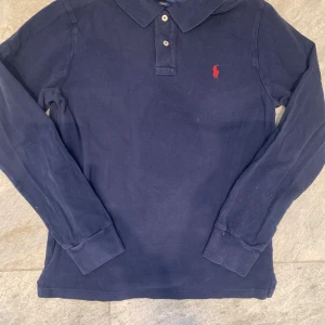 Mörkblå pikétröja från Polo Ralph Lauren - Snygg mörkblå långärmad pikétröja från Polo Ralph Lauren i slim fit-modell. Tröjan har klassisk krage, knäppning med två knappar och den ikoniska röda loggan broderad på bröstet. Tillverkad i mjuk bomull som är skön mot huden.