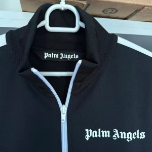 Palm Angels svart track tröja - Svart track jacket från Palm Angels med vita ränder längs ärmarna och vit dragkedja. Jackan har hög krage, ribbade muddar och logga på bröstet. Perfekt för en sportig och trendig look. 