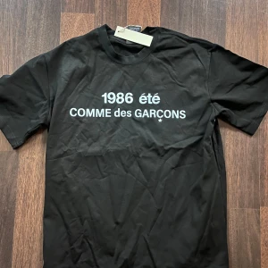 Svart Comme des Garçons t-shirt - Svart t-shirt från Comme des Garçons med vit text '1986 été' och logga på bröstet. Klassisk passform med rund hals och korta ärmar. Tillverkad i mjuk bomull, perfekt för en clean och stilren look.