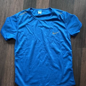 Blå Nike dri-fit t shirt - Snygg blå t-shirt från Nike med klassisk logga på bröstet och reflekterande detaljer på axlarna. Tillverkad i ett lätt och ventilerande funktionsmaterial som passar perfekt för träning. Kortärmad och med rund halsringning för en sportig look. Står M på tröjan men skulle snarare säga att det är tight S / loose XS